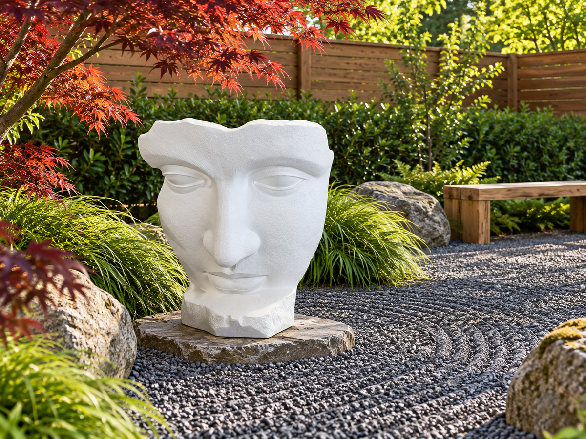 Sculpture de visage Atoll blanche dans une cour de gravier entourée de végétaux